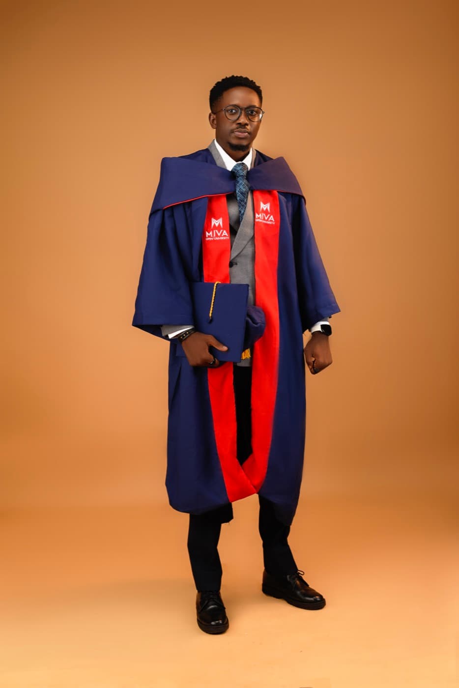 Blessing Ajala standing in Miva Open University graduation regalia, mortarboard tucked under one arm